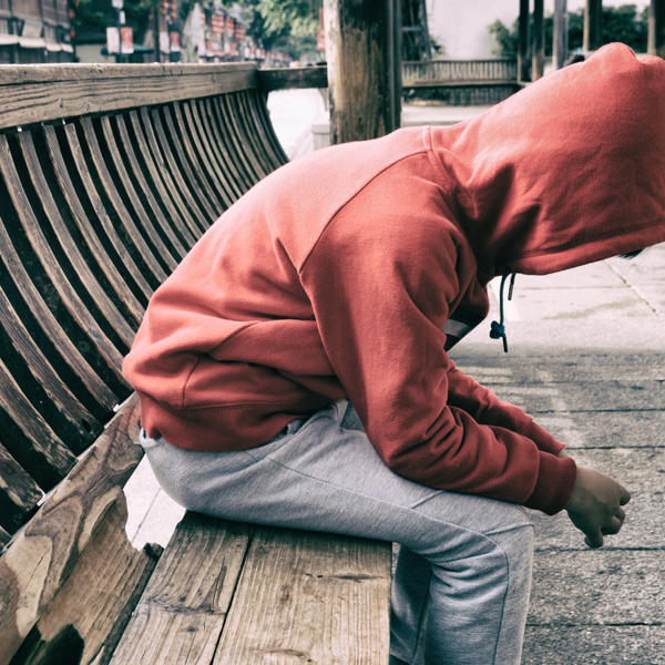 A photo of a man on a bench slouching forward with his hood up holding both of his hands together 